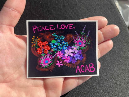 Colorful sticker with flowers over rusting handcuffs and Peace, Love, ACAB text held by a hand on a dark surface