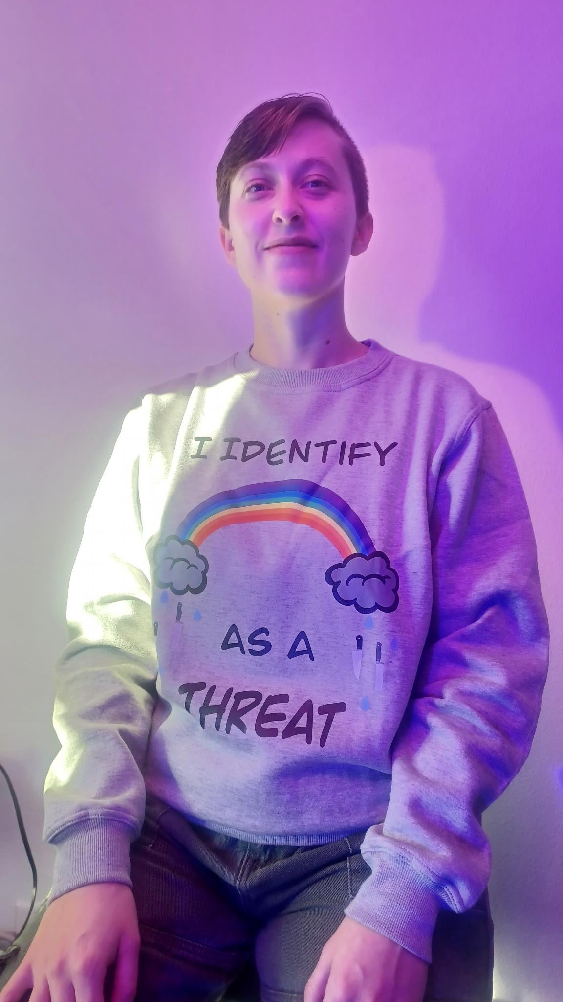KC wearing a jersey gray crewneck sweater with the words "I identify as a threat" with a rainbow with clouds raining knives