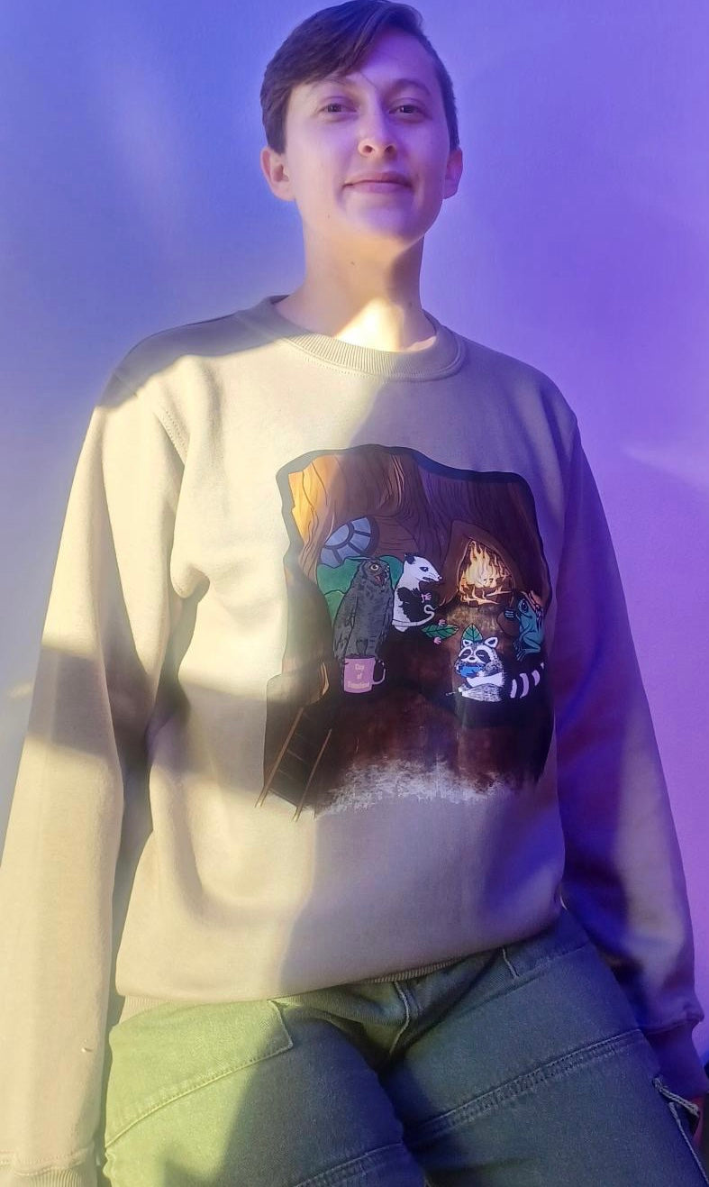 KC in light brown crewneck sweater with an image of an owl, possum, raccoon, and frog in a treehouse around a fire place all holding coffee mugs, the owl's coffee mug says "cup O sunshine"