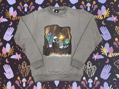 light brown crewneck sweater with an image of an owl, possum, raccoon, and frog in a treehouse around a fire place all holding coffee mugs, the owl's coffee mug says "cup O sunshine"